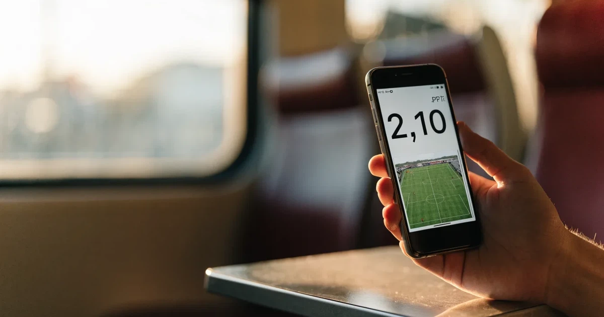 Uma mão a segurar um telemóvel com uma aposta no ecrã, num comboio ao fim da tarde